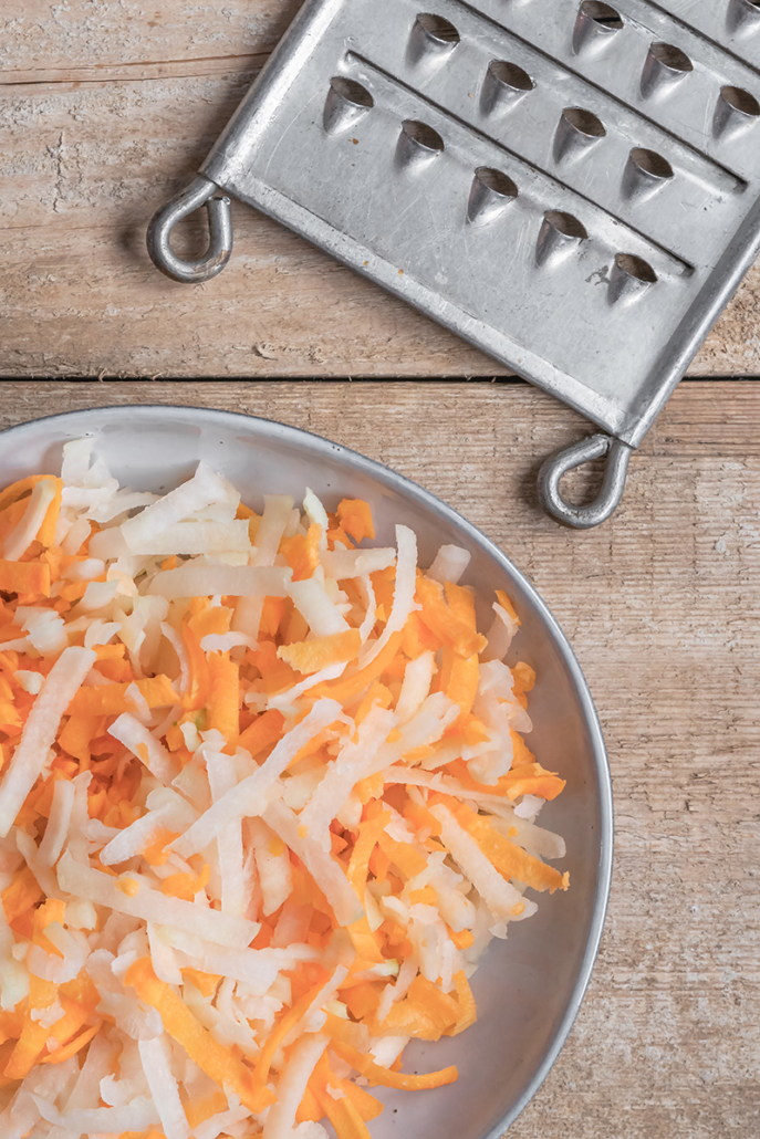 Geraspelte Karotten und Kohlrabi zusammen in einer Schüssel, daneben liegt eine Reibe