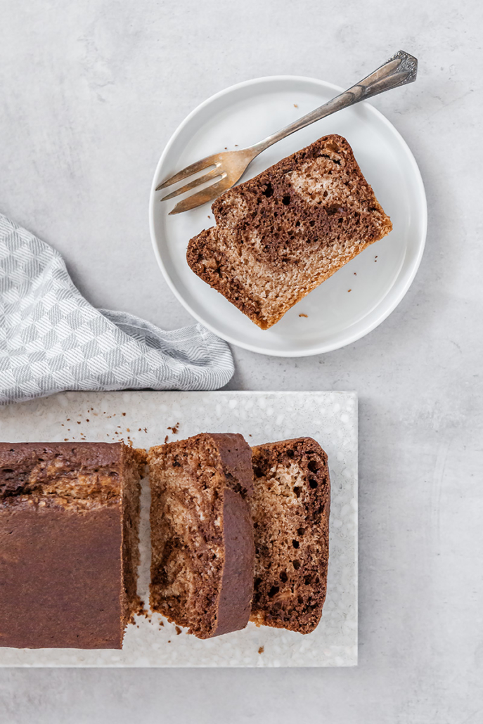 Angeschnittener Marmorkuchen auf einem Schneidebrett, daneben steht ein Teller mit einem einzelnen Stück Marmorkuchen und einer Kuchengabel.