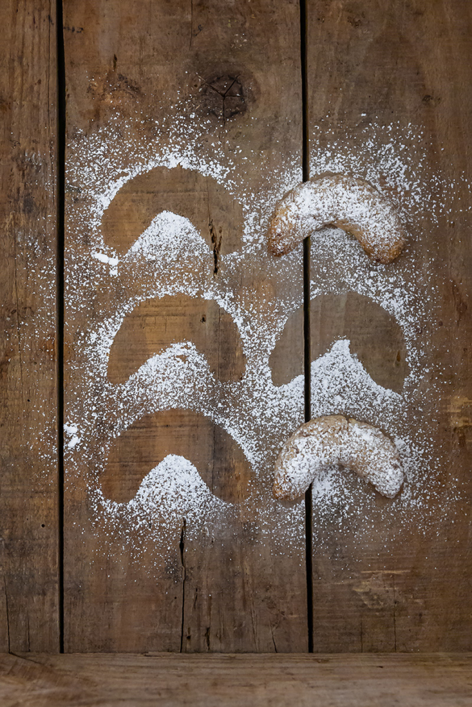 Mit Puderzucker betreute Haferflocken-Mandel-Kipferl auf einem alten Holzuntergrund. Vier von sechs fehlen und nur die Abdrücke im Puderzucker sind zu sehen.