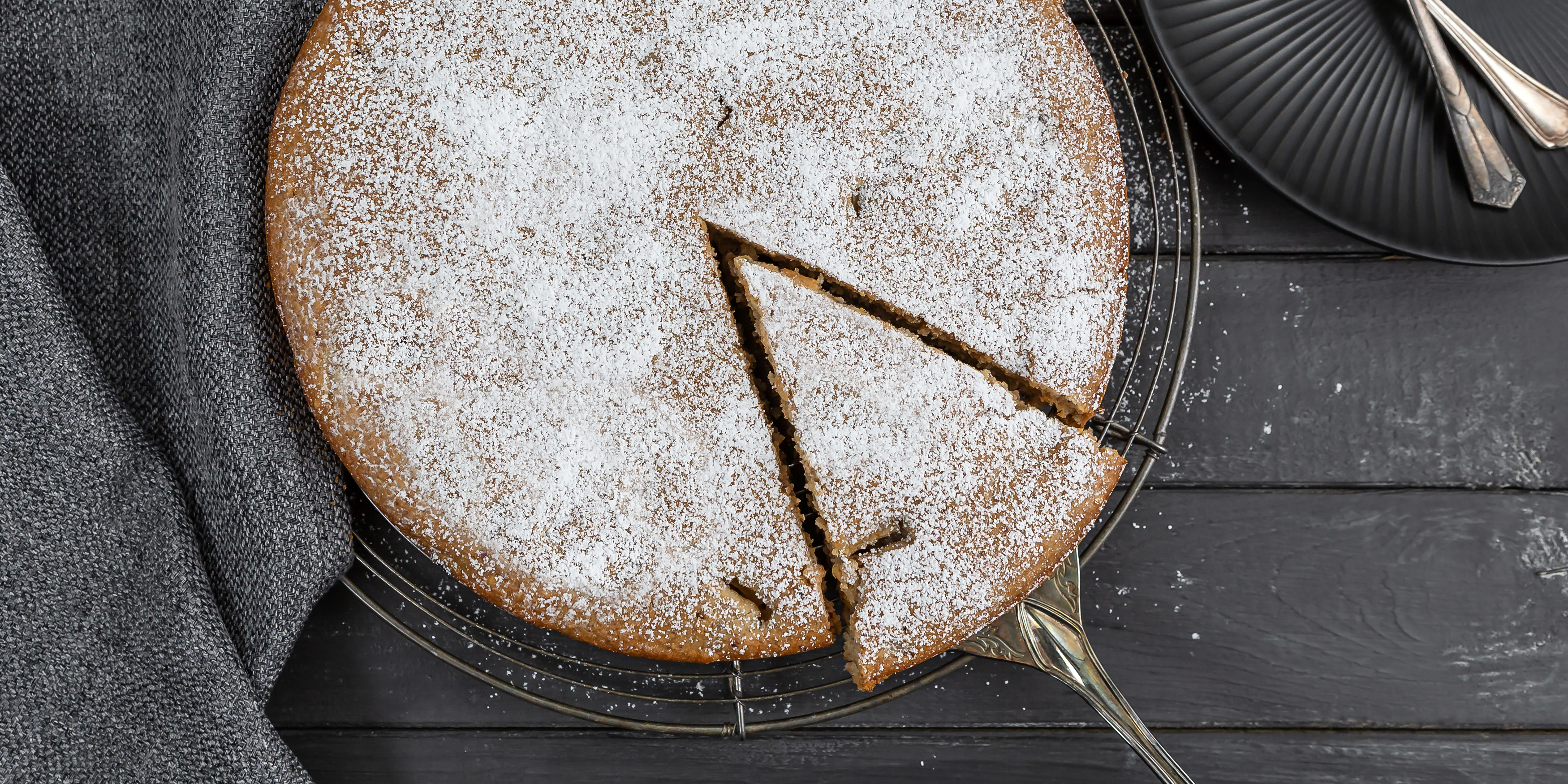 Angeschnittener Rührkuchen mit Äpfeln und Zimt auf einem Kuchengitter. Ein Stück wird mit dem Kuchenheber herausgezogen. Ein Teller mit zwei Kuchengabeln steht bereit.